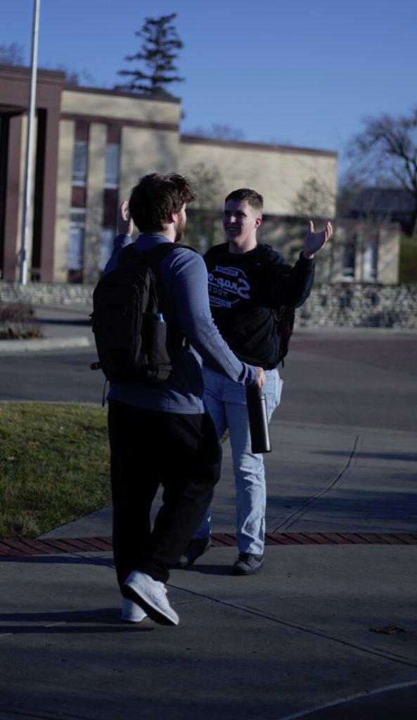 Two people with backpacks greet each other energetically on a sidewalk in front of a building. One person holds a bottle while gesturing with his arms.